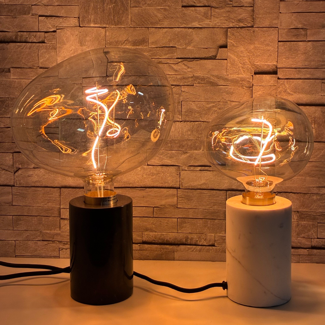 Två asymmetriska LED-glödlampor i skimrande glas på svarta och vita marmorfötter. Glödlampor med skimrande glas och spiralformad LED-tråd – stämningsfull belysning mot stenvägg.